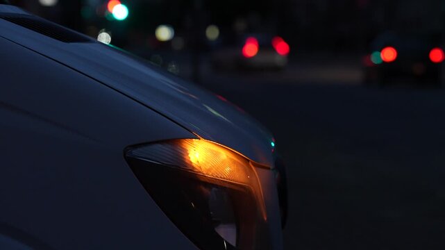 Car front indicator glowing amber beside a headlamp as traffic passes along a city street, showing vehicle signaling equipment used in urban road traffic circulation