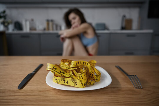 Depressed anorexic woman looking at plate with measuring tape served for dinner