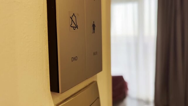 Hotel room status panel next to doorway shows muted bell icon, DND letters, and small marker on beige wall under warm interior lighting, with hotel, room, status, panel detail