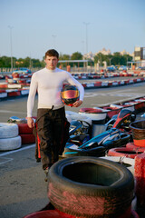 A confident young kart racer holding a helmet stands at a vibrant outdoor karting track © Nomad_Soul