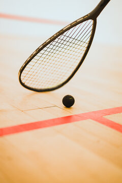 Squash racket and black ball positioned on an indoor wooden squash court with red lines.