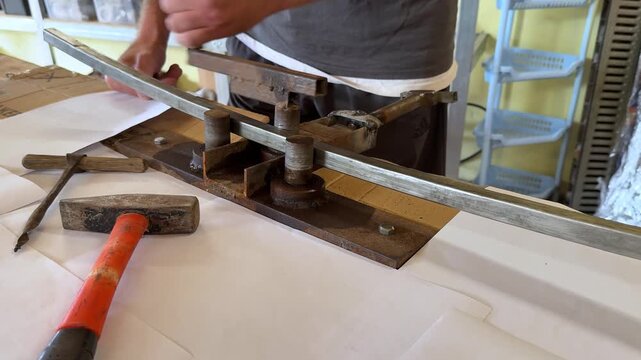 Skilled worker using metal bending tool to shape a metal strip on a workbench, with tools and materials visible in a workshop environment