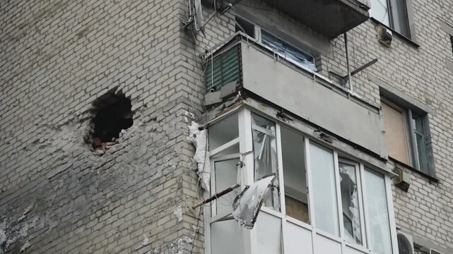 Damaged urban apartment building facade with large wall opening, collapsed balcony, shattered glass windows, scattered debris, and exposed gray masonry after collapse