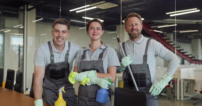 Cleaners team of janitors in office cleaning. Smiling staff in uniforms wear gloves holding spray bottles and a mop by a meeting table. Clean team symbol of workplace hygiene and trust.