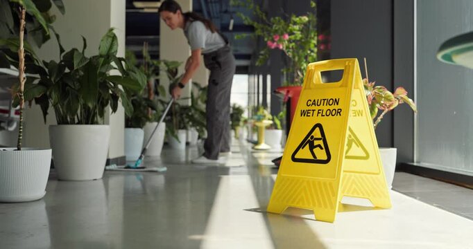 Cleaner mopping office floor near safety sign. A professional janitor in uniform cleans a bright corridor with potted plants, promoting hygiene and slip risk prevention. Key theme is safe work.