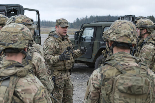 Group of soldiers gathering near military vehicle in muddy field, camouflage uniform troops briefing outdoor with forest background, tactical mission concept