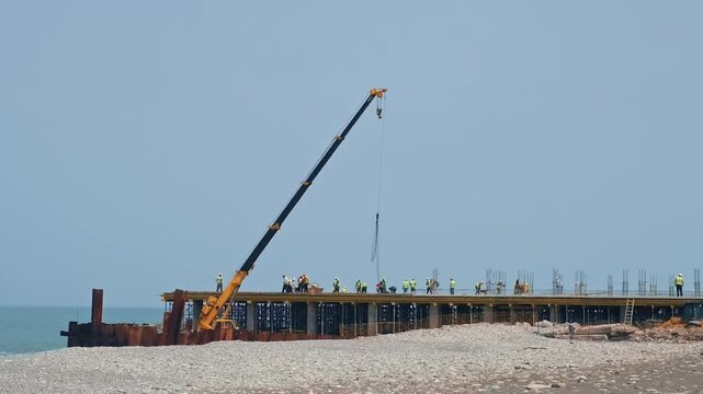 Coastal pier construction as crane boom lowers hoist cable and hook over exposed rebar while workers in high-visibility vests and hard hats gather on deck beside shoreline