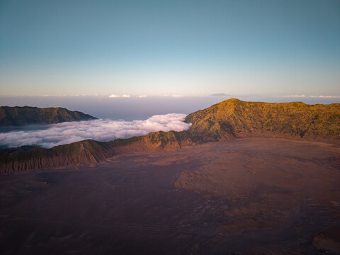 Sunlight bathes rugged volcanic landscape. Clouds roll below jagged mountain peaks at Bromo