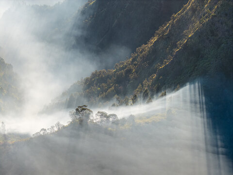 Mist rolls through rugged mountains, soft and slow. Sunlight breaks through clouds at Sakapura near Bromo
