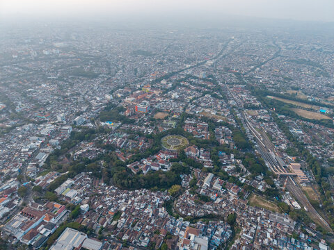 Aerial panorama of dense Malang neighborhoods, rooftops, roads, railway lines, green spaces, and urban development under hazy skies.
