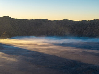 Sunlight glows over misty, volcanic valley. Clouds drift gently across barren, rugged terrain at Bromo © Cloudyew