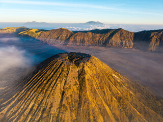 Aerial view of Gunung Batok rugged volcanic slopes, ash plain, surrounding peaks, and drifting clouds in golden light. © Cloudyew