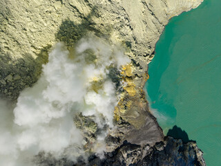 Volcanic smoke billows from rugged, rocky terrain. Turquoise water laps against the cliffside edge at Mount Ijen © Cloudyew