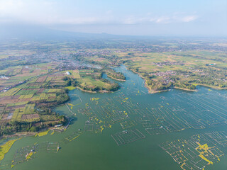 Drone panorama of Karangkates Reservoir, floating fish cages, cultivated fields, villages, and hazy mountain landscapes in Indonesia. © Cloudyew