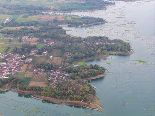 Drone panorama shows floating fish cages, cultivated fields, forested shoreline, and rural villages around Karangkates Reservoir in Indonesia. © Cloudyew