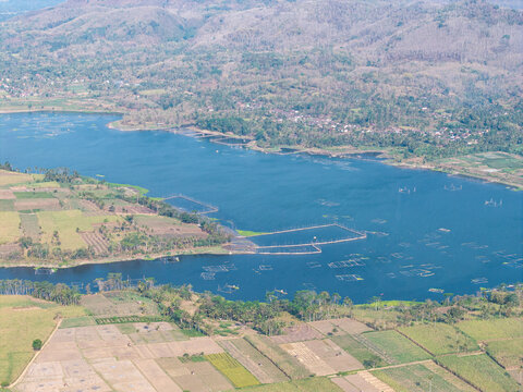 Drone panorama of Waduk Karangkates reservoir with floating fish farms, tropical villages, cultivated fields, palms, and forested hills.