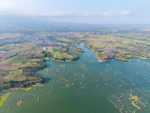 Drone panorama of Karangkates Reservoir, floating fish cages, cultivated fields, villages, and hazy mountain landscapes in Indonesia.