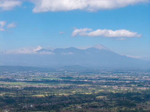 Vast valley stretches beneath towering mountain peaks. Clouds drift lazily across bright blue skies at Malang. Back is Mount Sermeru