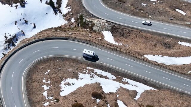camper van winds through tight s curve second car trails above julier pass mountain road patchy snow dry terrain guardrail graubunden switzerland drone aerial view high altitude road trip
