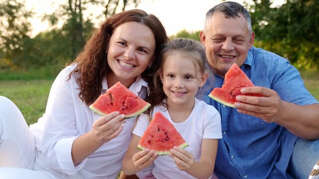 Happy mother, father, daughter playfully hide their faces behind slices of watermelon before lowering them to smile and take big bite. Summer picnic and healthy family lifestyle concepts