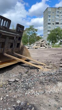 Heavy machinery bucket with forks positioned on construction site, surrounded by debris and a residential building in the background under a blue sky with clouds