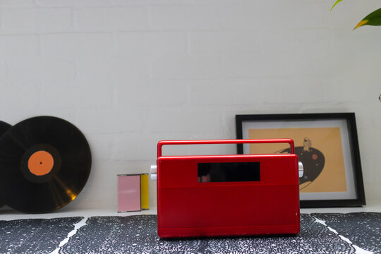 Red portable radio is sitting on patterned cloth with records, cassettes, framed print, plant leaf