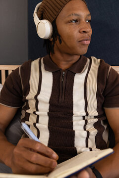 Male sitting on bed wearing brown beanie and white headphones, holding pen and writing in notebook