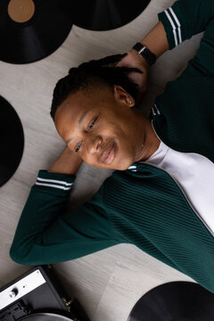 Adult African male lying on light wood floor amid vinyl records and turntable, wearing green jacket