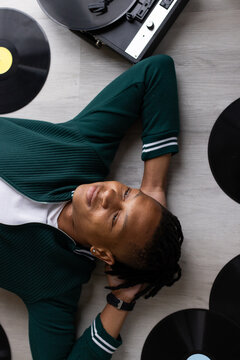Male lying on light floor wearing green jacket watch vinyl records surrounding him and turntable