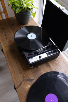 Portable turntable is sitting open on wooden table by window, vinyl and plant basking in daylight