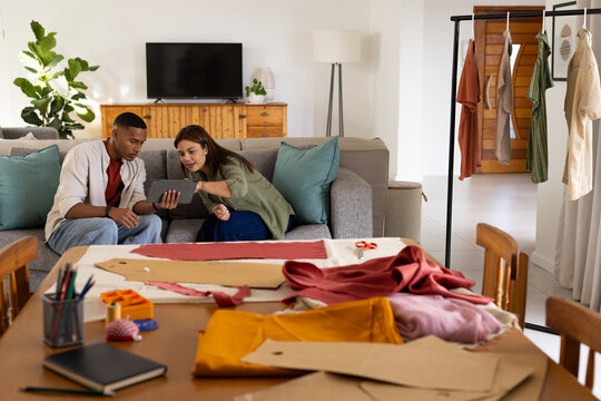 African American couple sitting on gray sofa, holding tablet and arranging fabric patterns on table