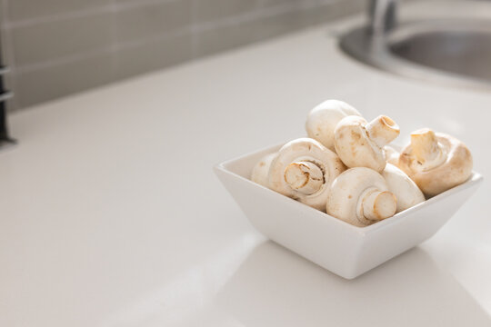 White square ceramic bowl is sitting on light counter near sink rim, showing mushrooms, copy space
