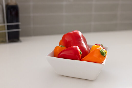 White square ceramic bowl is holding red and orange bell peppers on white countertop
