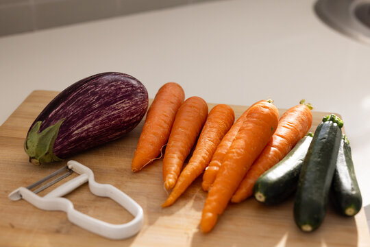 Wooden cutting board is holding striped eggplant, carrots, zucchinis, peeler on counter near sink