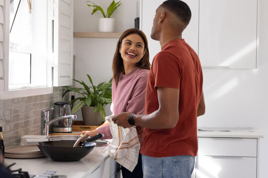 African American couple standing at kitchen sink holding frying pan and spatula, man holding towel
