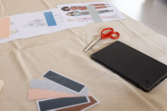 Red-handled scissors are resting above black tablet on beige fabric work surface, with swatch cards