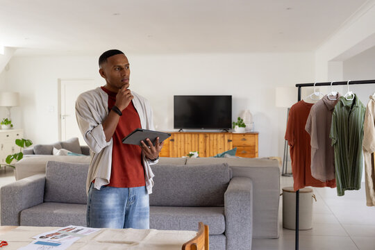 African American man standing holding tablet and touching chin in living room with clothing rack