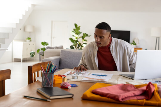 African American man sitting at wooden table sketching designs and working on silver laptop at home