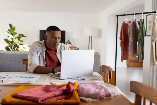 African man working on silver laptop at dining table with fabric swatches, wearing rust-red shirt
