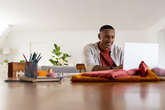 African American man working on laptop at dining table with colored pencils, wearing rust tee
