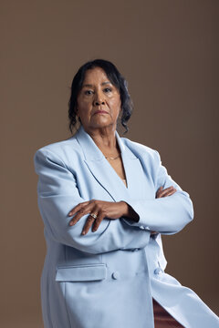 Senior African American woman sitting in studio, wearing blue blazer, gold necklace, rings, gazing