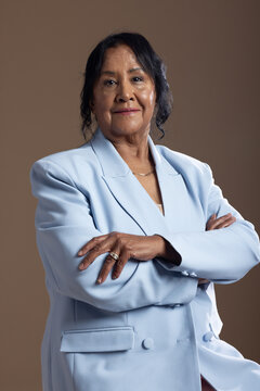 Senior African American woman posing with arms crossed in studio on brown backdrop wearing blazer