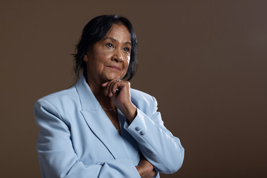 Senior African American woman posing, holding chin, wearing blue blazer, necklace, ring in studio