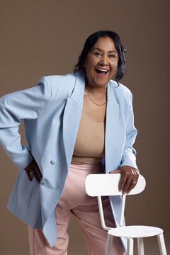 African American senior woman laughing and leaning on white chair in studio wearing blue blazer