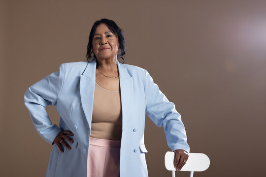 Senior African American woman standing in studio, hand on white chair, wearing light blue blazer