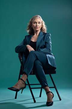Senior woman posing, looking toward camera on black metal folding chair in studio wearing teal suit