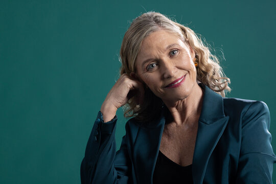 Senior woman sitting in studio on teal backdrop wearing teal blazer black top yellow stud earring