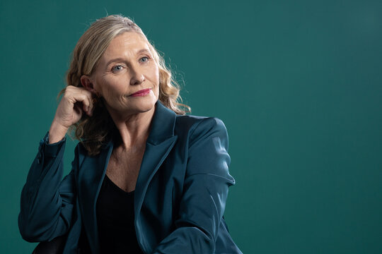 Senior female sitting and posing in studio, wearing teal blazer, black top, small hoop earring