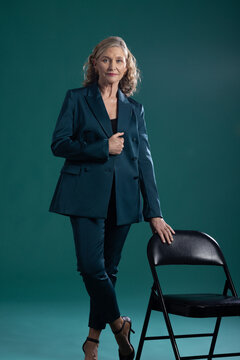 Senior woman wearing suit, heels, standing in studio, touching lapel, hand resting on folding chair