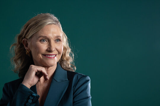 Senior female posing in studio, wearing teal blazer and gold hoops, resting fingers near chin